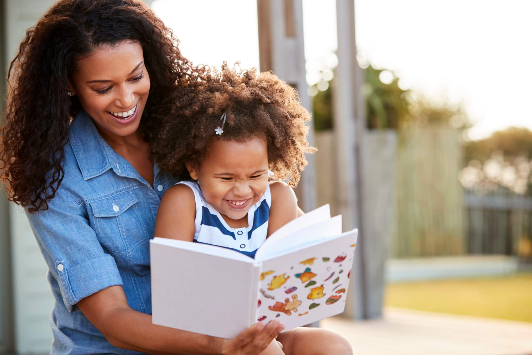 Young black girl reading book sitting on mums knee outdoors